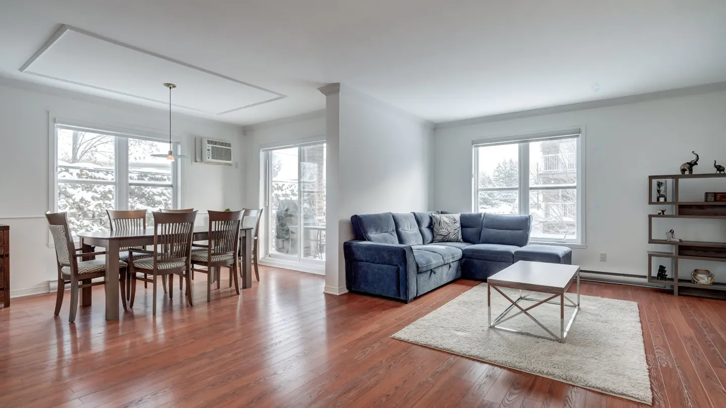 Bright living and dining area with hardwood floors, white walls, and large windows. A blue sectional sofa and dining table with chairs are visible.