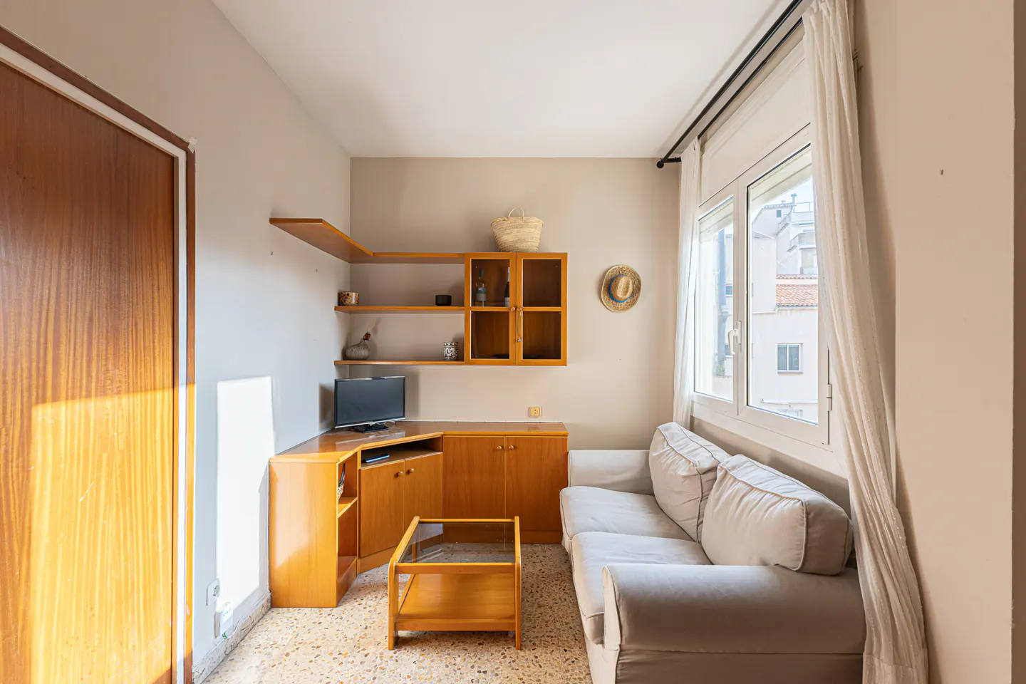 A cozy living room with a beige sofa, wooden furniture, and a window with white curtains. A TV sits on a corner cabinet, and shelves display decorative items.