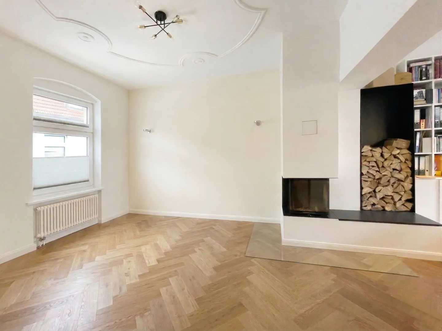 Bright living room with herringbone wood floors, white walls, and a modern fireplace with stacked firewood. A window with blinds and a radiator are visible.