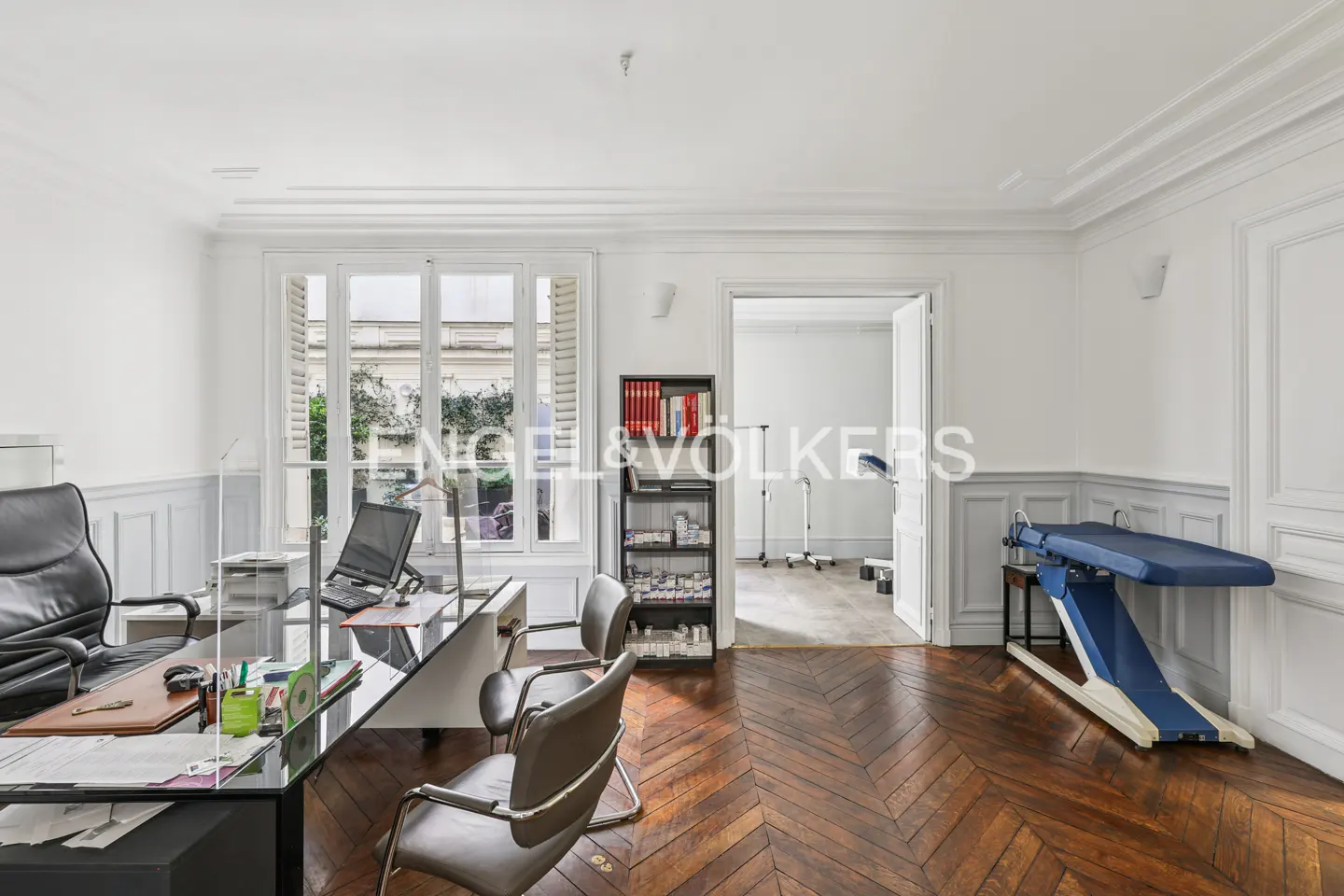 A doctor's office with a desk, computer, chairs, and a medical bed in a white room with wood floors.