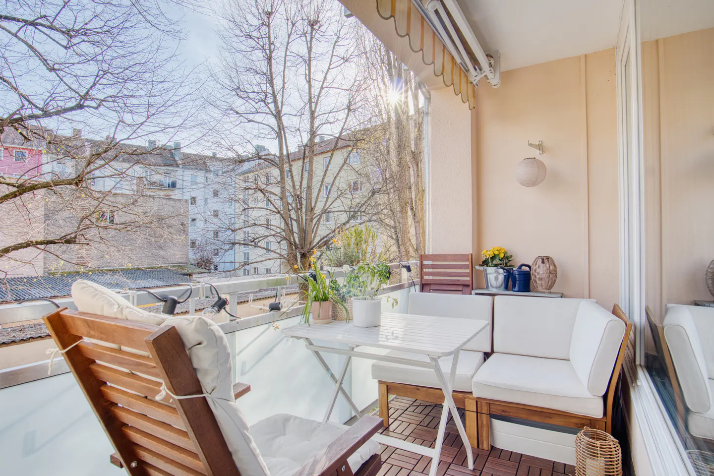 A sunlit balcony with a wood floor, white cushions, a table, and a view of buildings and trees.