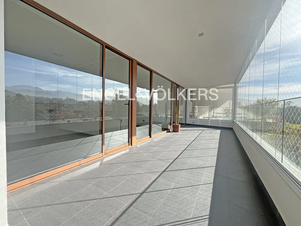 A long, covered balcony with gray tile flooring, glass doors, and a wire mesh railing. Mountains are visible in the distance.