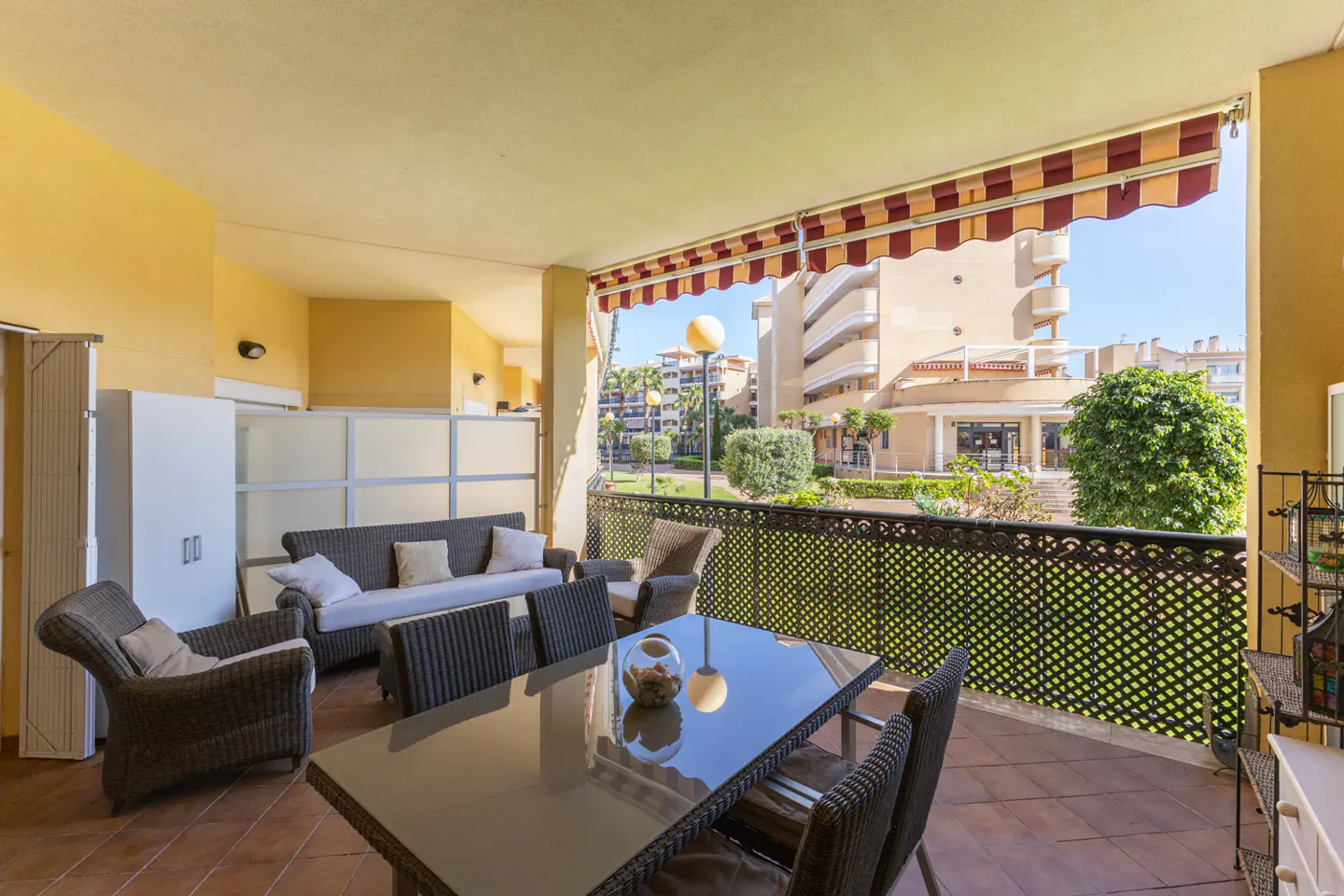 A sunny balcony with wicker furniture, a glass table, and a red and white striped awning overlooking a garden and buildings.