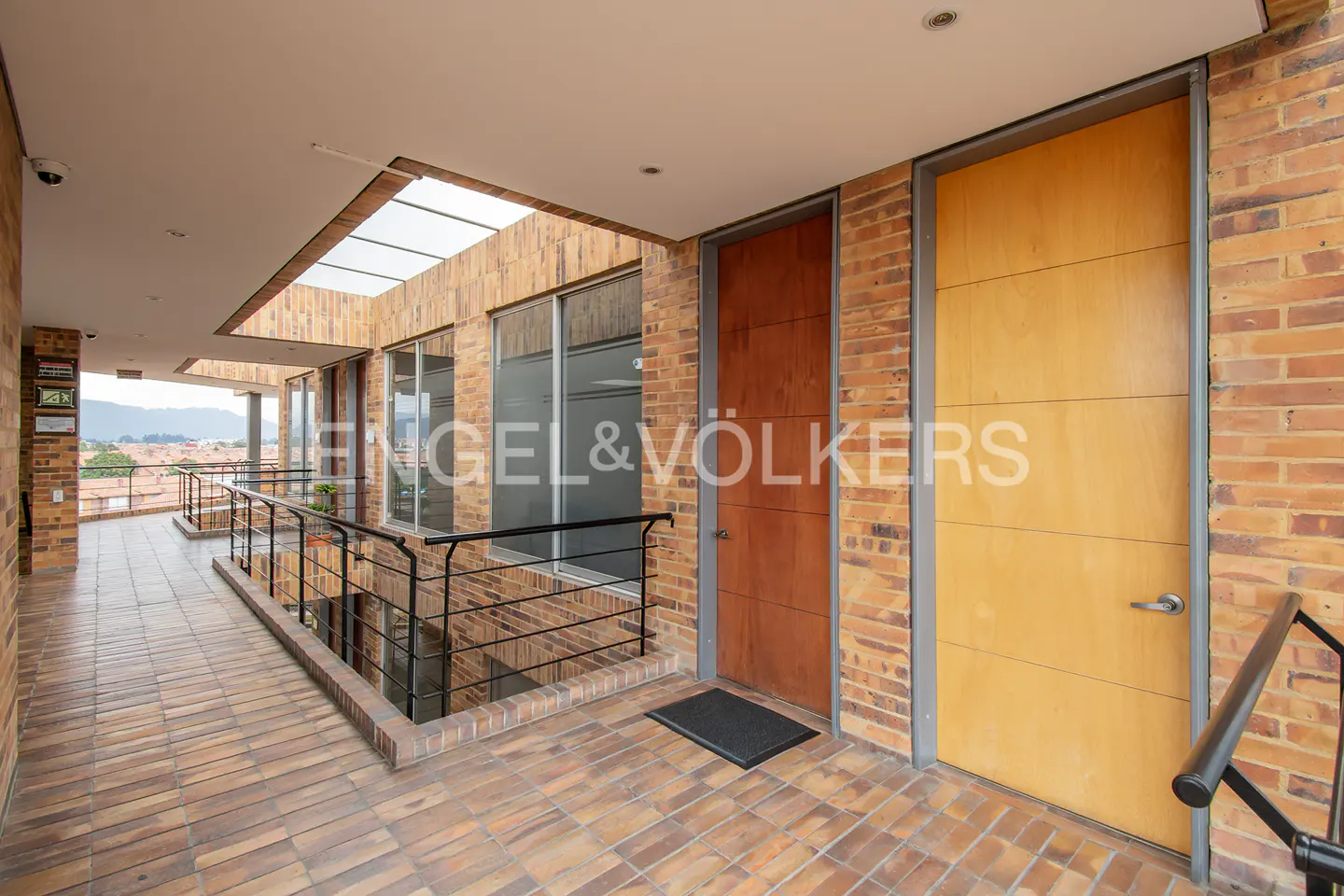 Hallway with brick walls and tiled floors, featuring two wooden doors and a black railing overlooking a cityscape.
