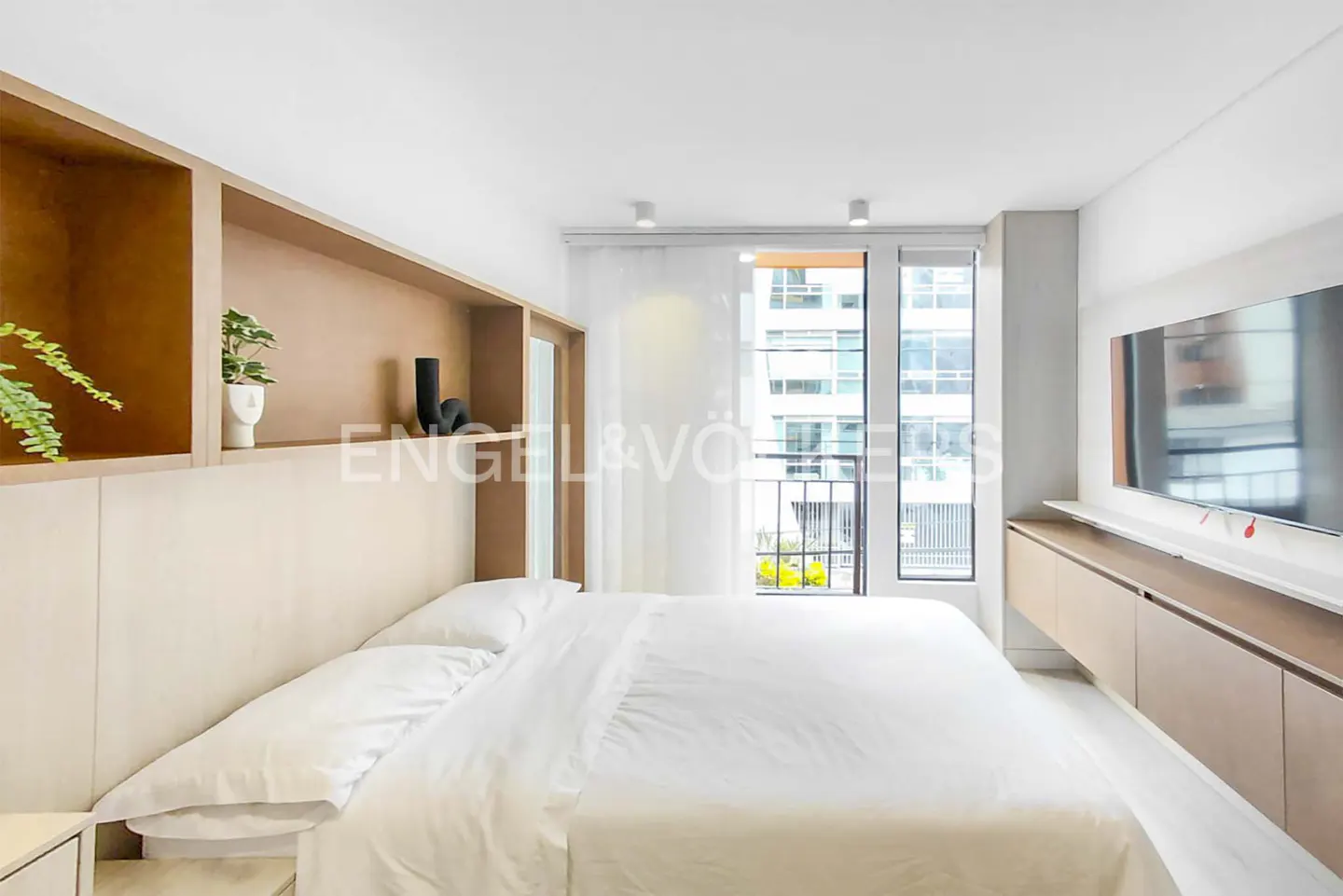 Modern bedroom with a white bed, light wood shelving, a wall-mounted TV, and a window overlooking a city building.