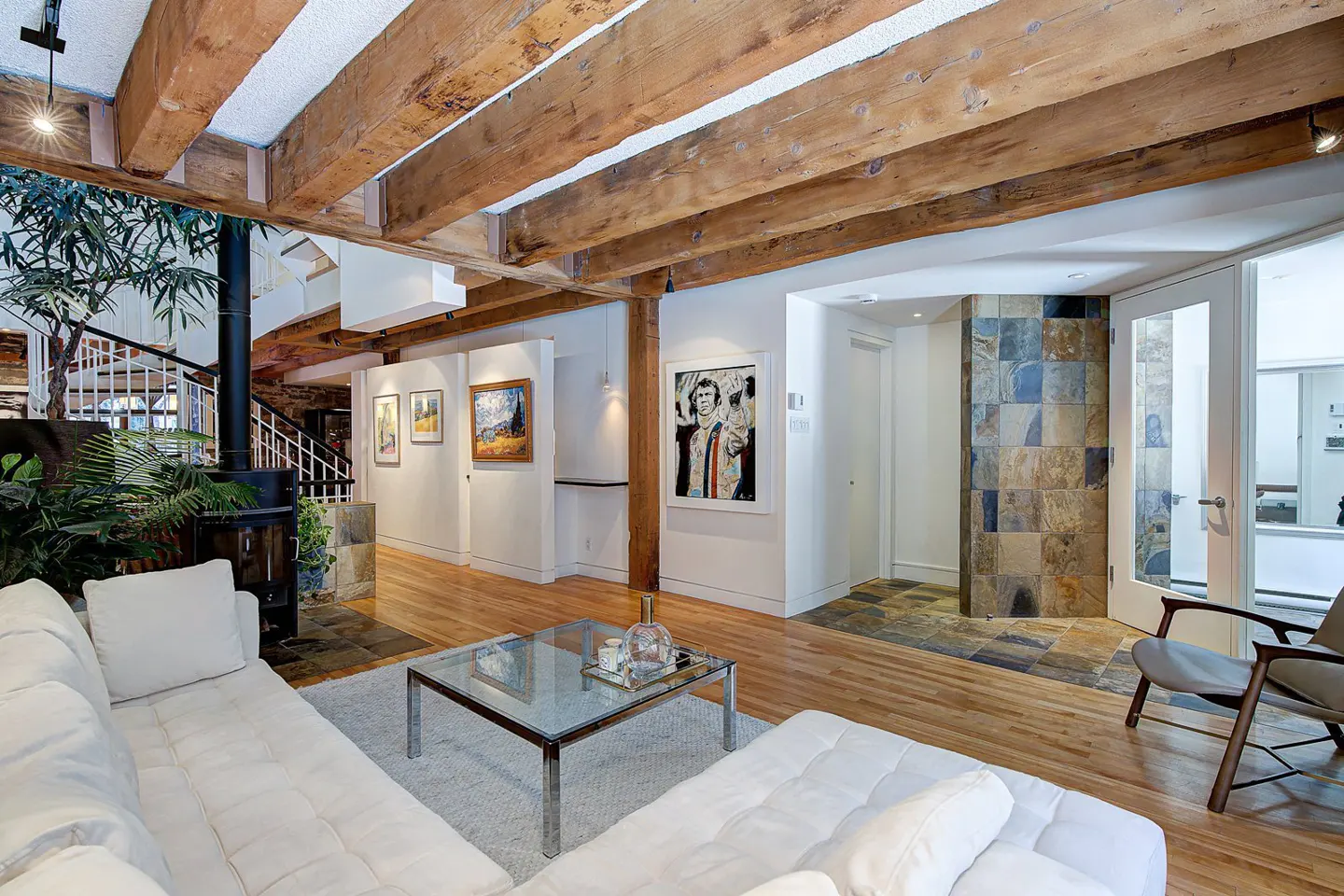 Living room with exposed wood beams, hardwood floors, and white sectional sofa. Art hangs on the white walls. A glass coffee table sits on a white rug.
