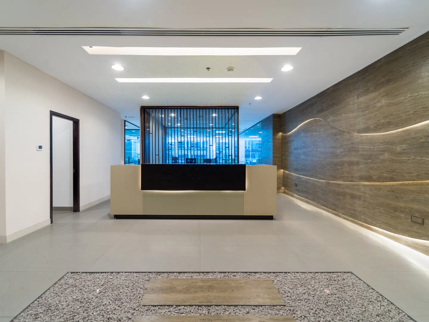Modern office lobby with a beige reception desk, black accents, and a decorative stone feature on the floor. A textured wall adds visual interest.