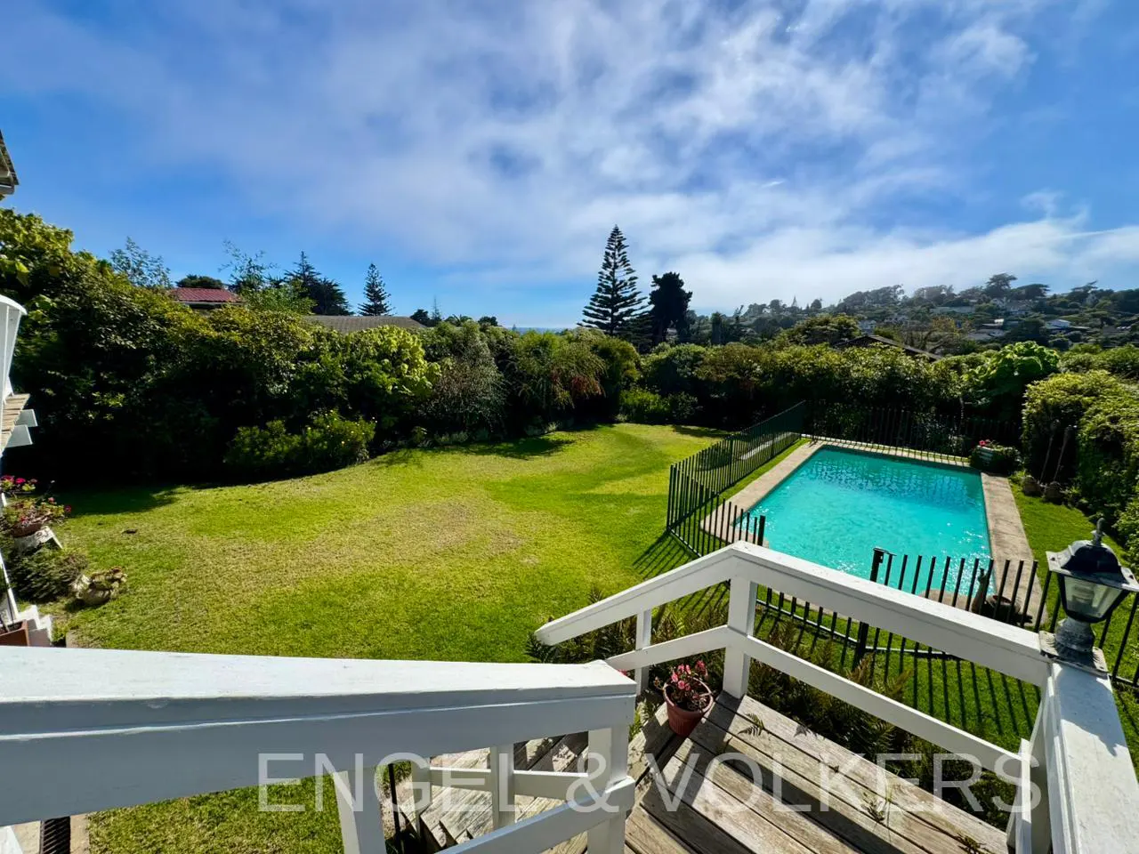 View from a wooden deck overlooking a green lawn and a turquoise swimming pool surrounded by lush trees under a blue sky.