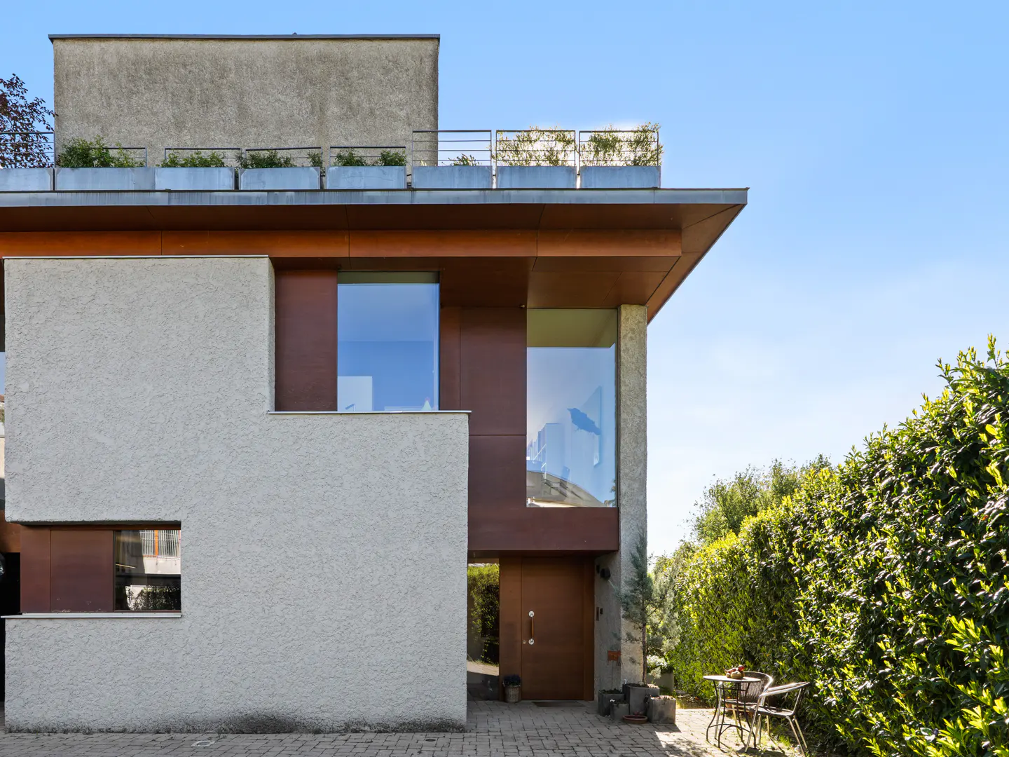 Modern home exterior with gray stucco and brown wood accents. Large windows reflect the blue sky. A rooftop garden adds greenery.