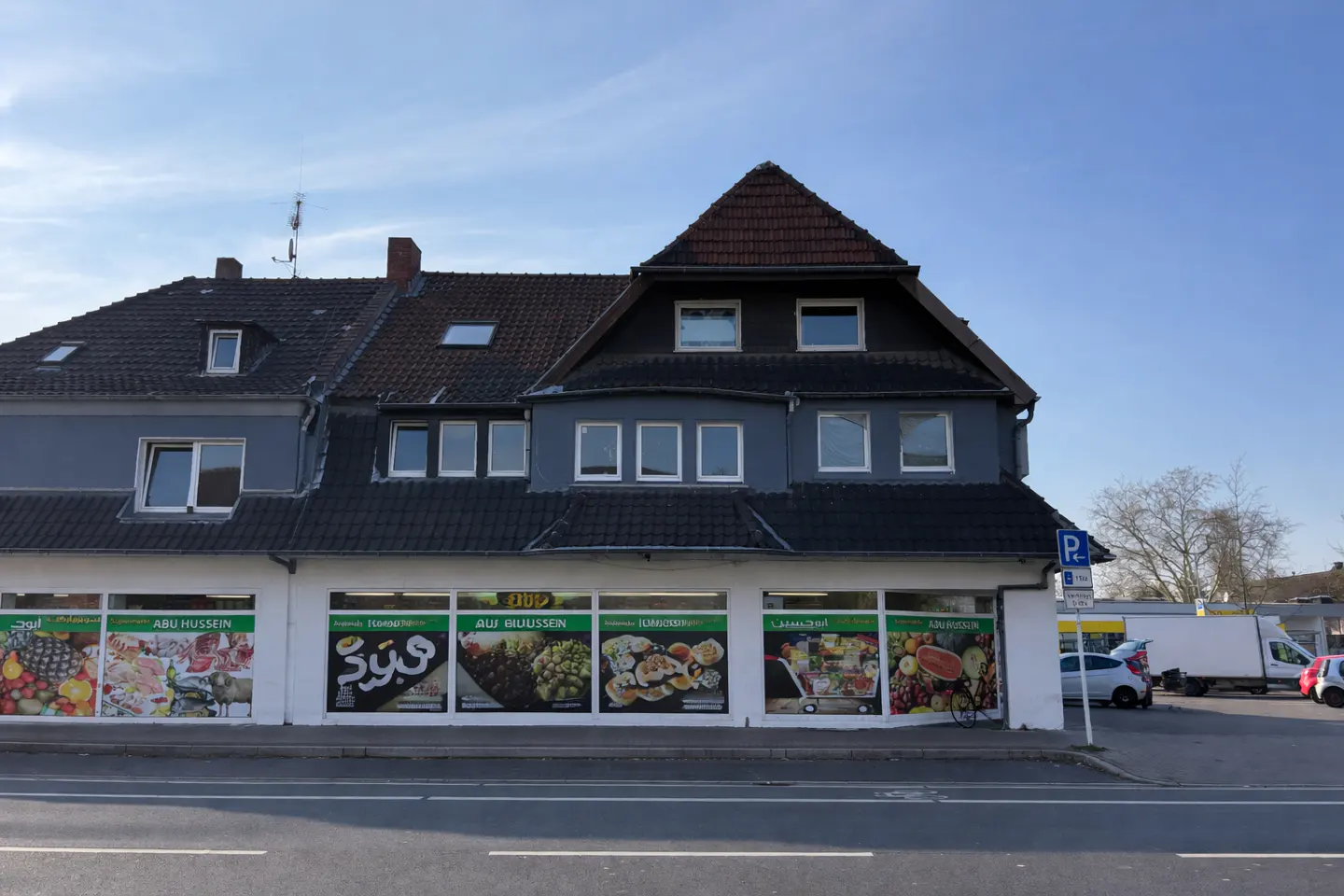 Two-story gray building with a dark roof. The ground floor has large windows with signs for "Abu Hussein" and food images. A parking sign is visible.
