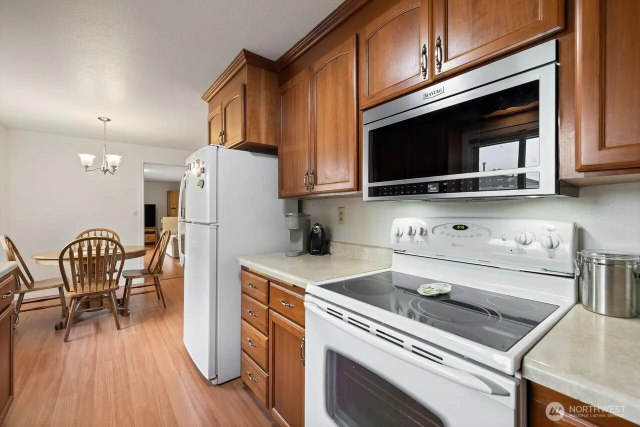 A kitchen with wood cabinets, white appliances, and a dining area. A white refrigerator stands next to the cabinets. A white stove and microwave are visible.