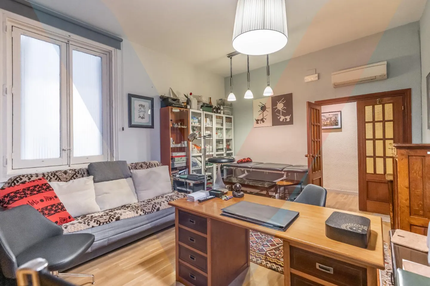 A bright office space with a wooden desk, sofa, massage table, and cabinets filled with supplies.