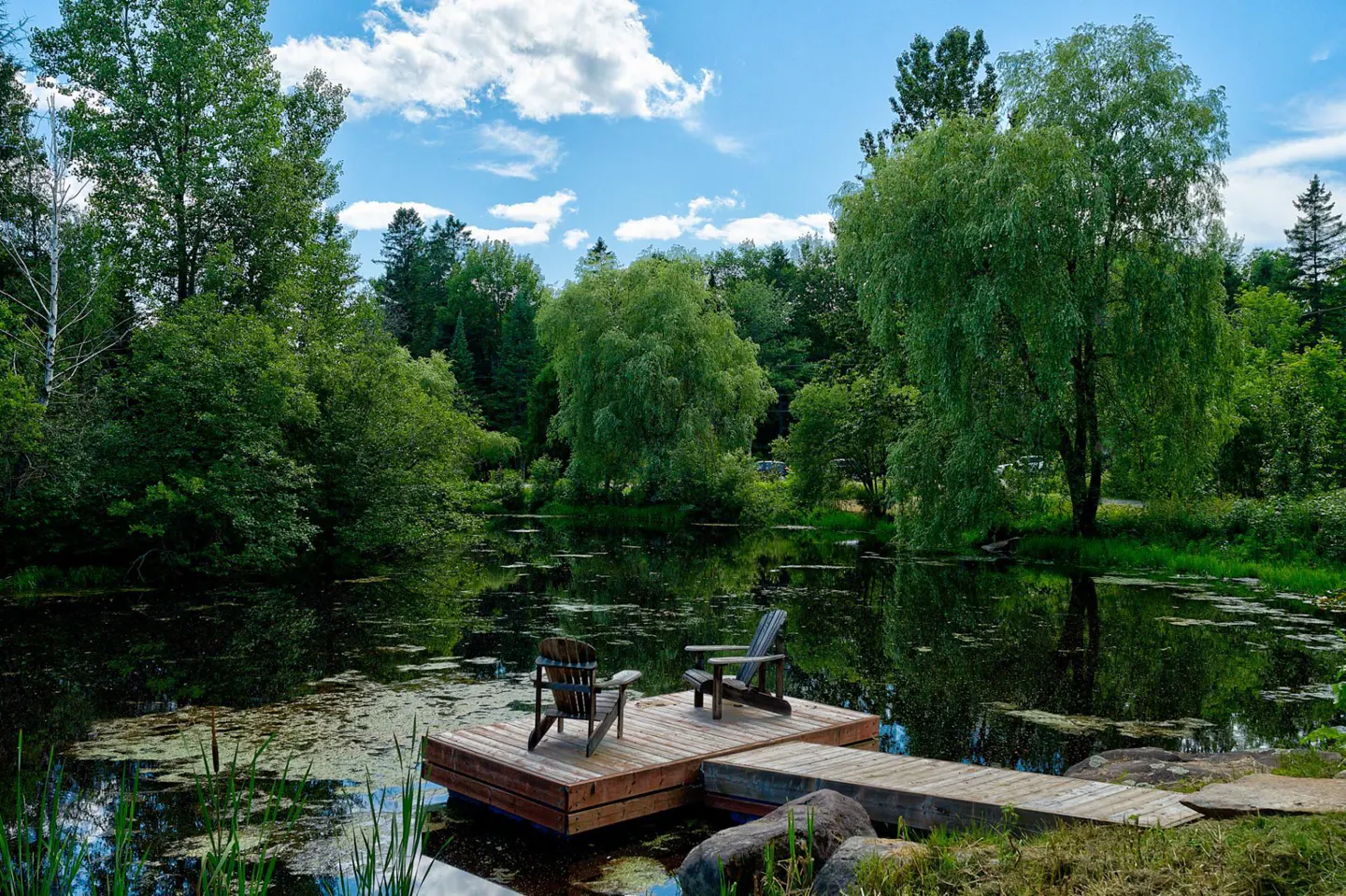 A tranquil pond with a wooden dock featuring two Adirondack chairs, surrounded by lush green trees under a partly cloudy sky.