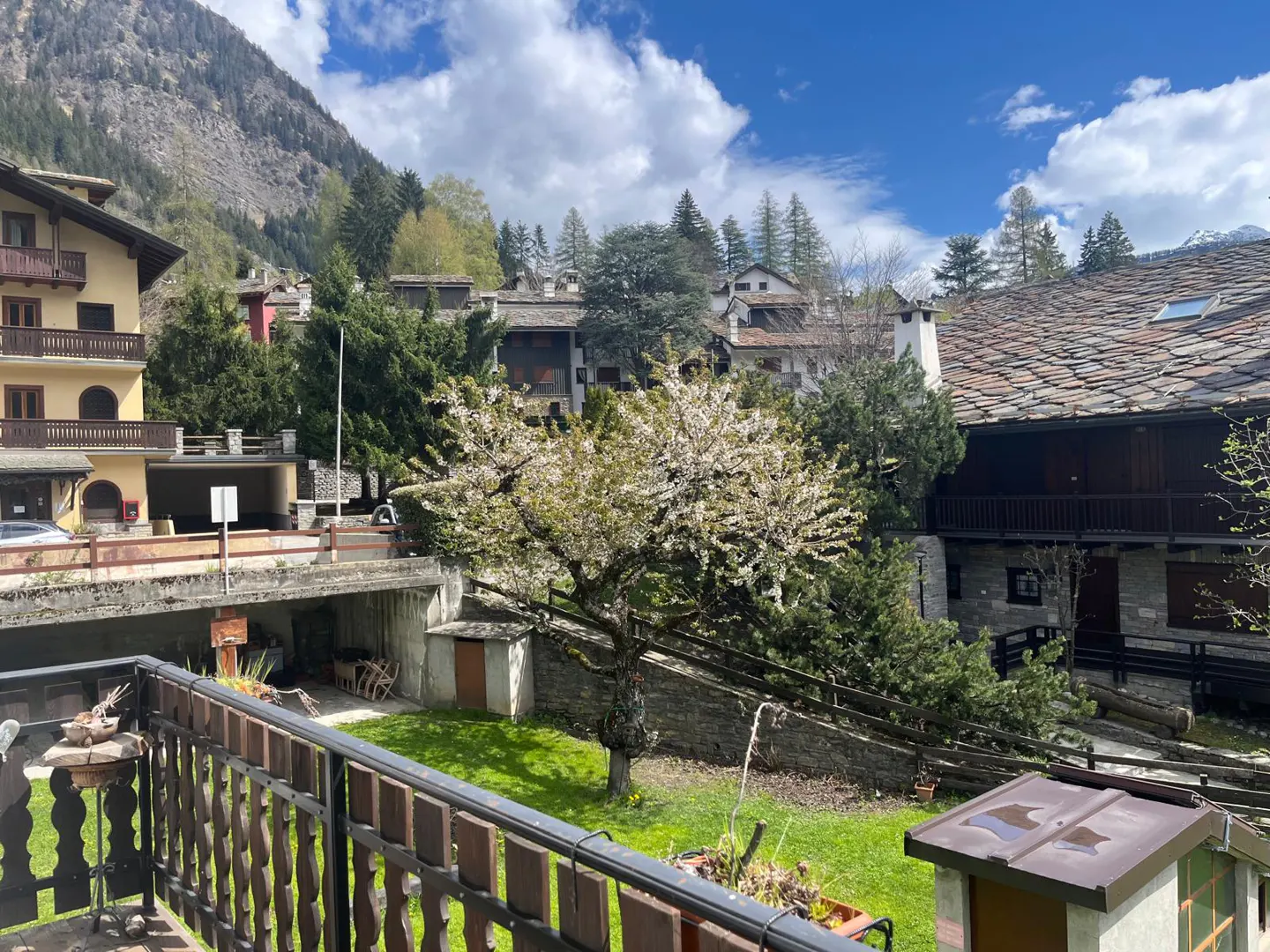 View from a balcony overlooking a green lawn, a flowering tree, and stone buildings in a mountain village under a blue sky.
