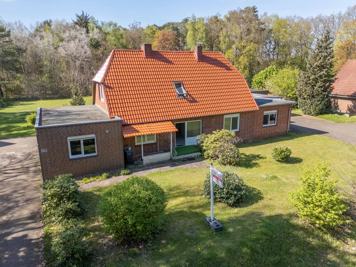 Aerial view of a brick house with an orange tile roof, green lawn, and a "For Sale" sign in the yard.
