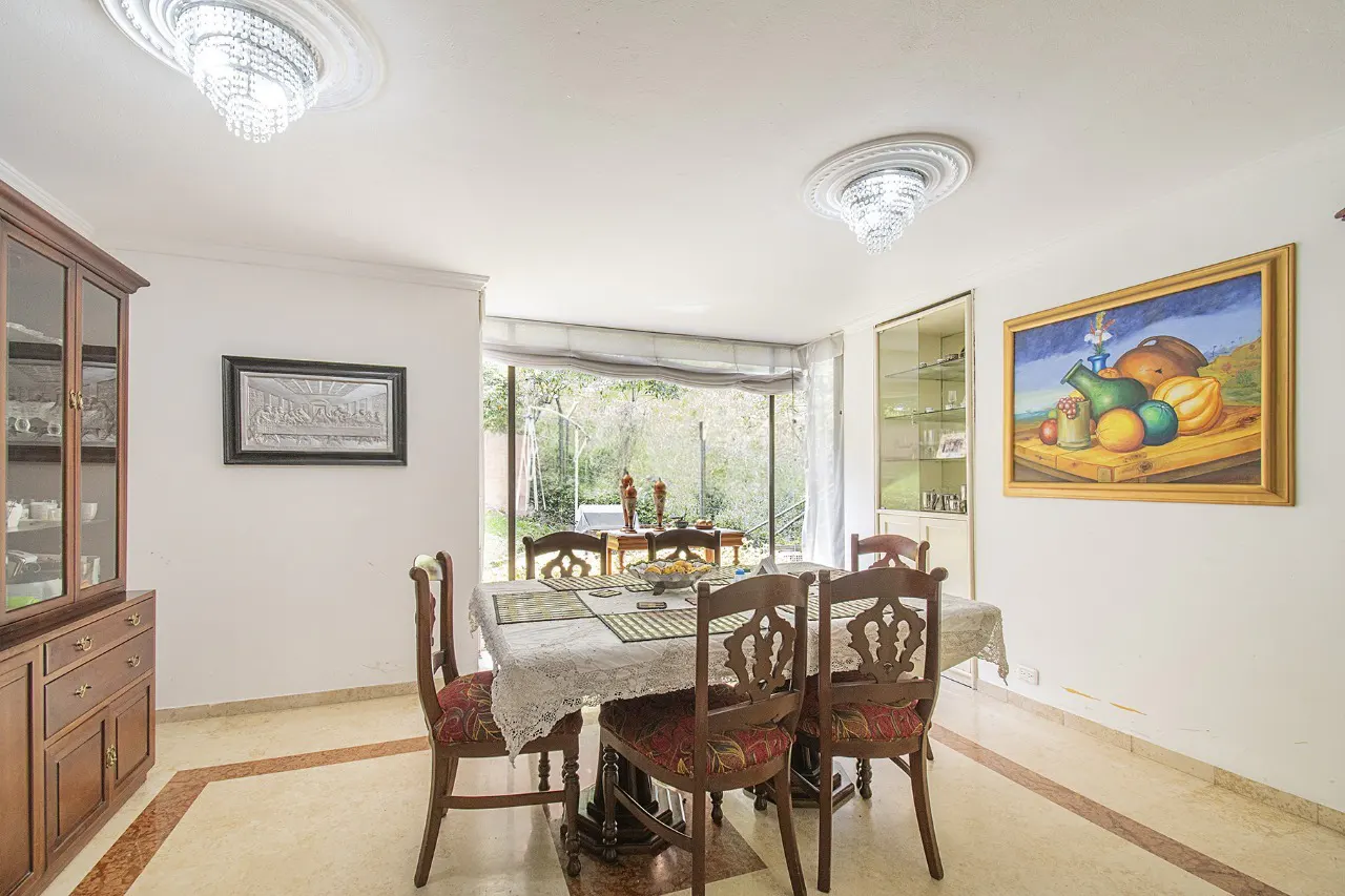 Bright dining room with a table, chairs, and a large window overlooking a green outdoor space. Artwork and a china cabinet add to the room's decor.