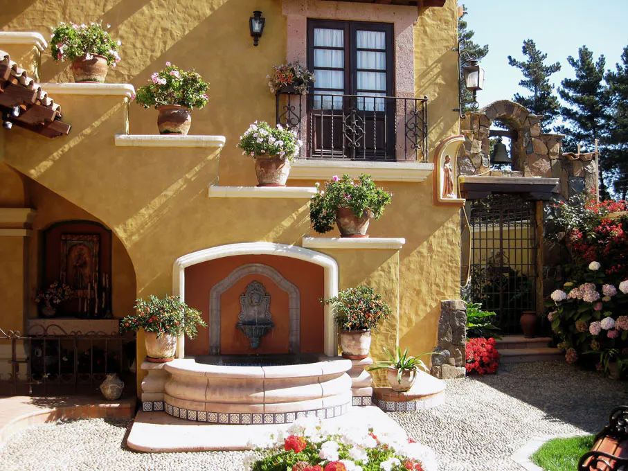 Exterior of a yellow stucco house with a fountain, flower pots, and a wrought iron balcony.