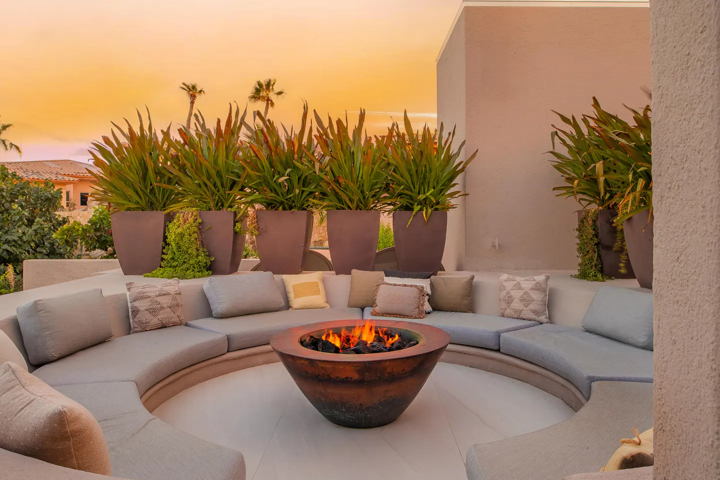 Outdoor patio with a circular sofa, pillows, and a fire pit. Tall potted plants line the back wall, with a sunset sky in the background.