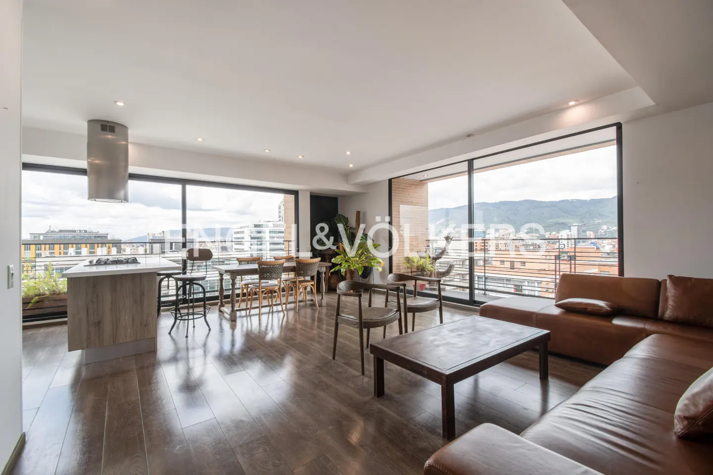 Modern open-plan apartment with a brown leather sofa, dining area, kitchen island, and floor-to-ceiling city view windows.