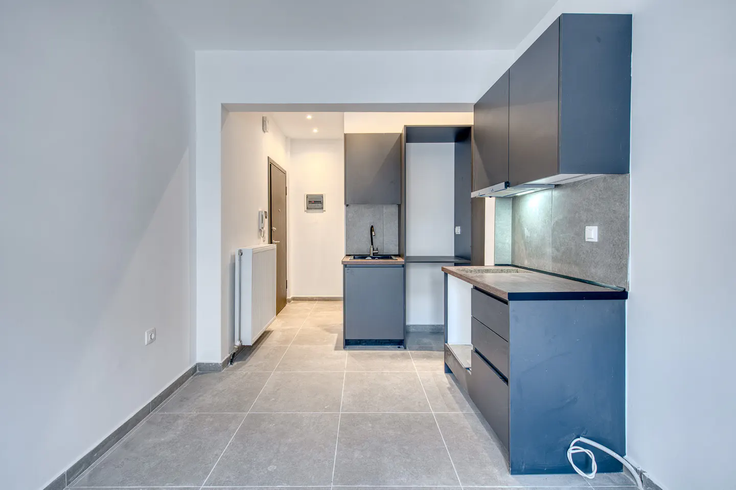 A modern apartment interior with gray tile flooring, white walls, and a compact gray kitchen area.