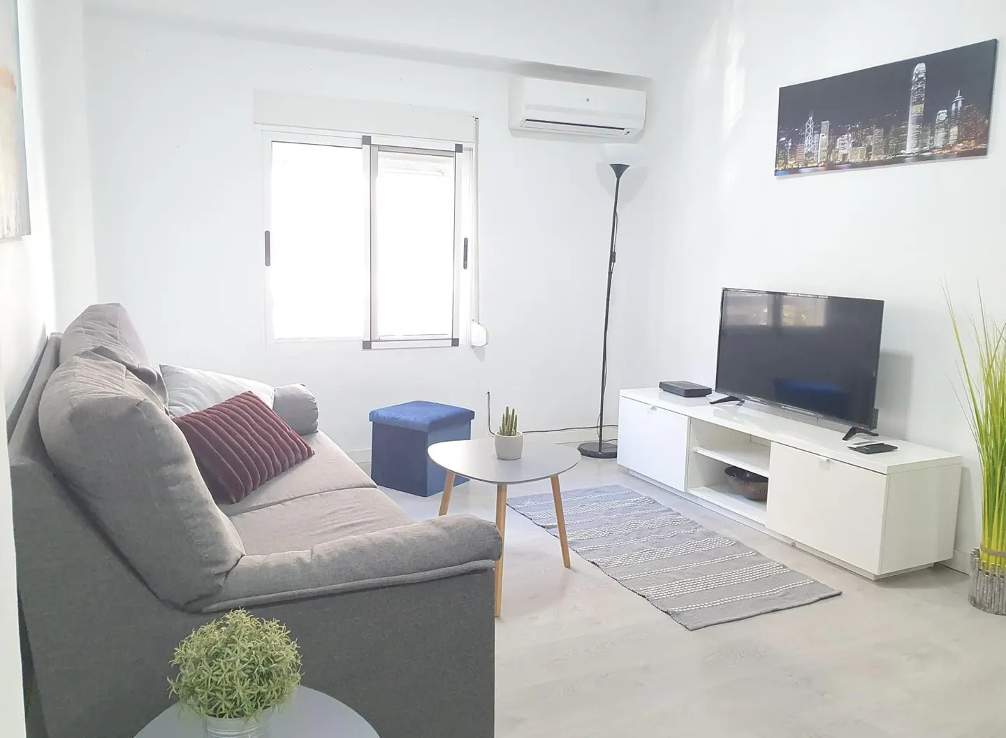 Bright living room with a grey sofa, white media console, flat-screen TV, round coffee table, and city skyline wall art.