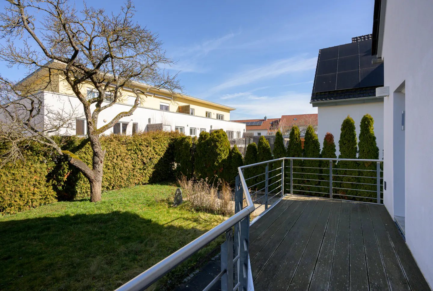 View from a wooden deck with metal railing, overlooking a green lawn, trees, and neighboring houses under a blue sky.