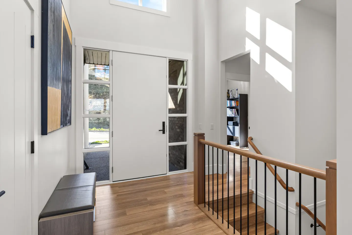 Bright, modern foyer with white walls, wood floors, and a white front door with glass panels. A gray bench sits near an abstract painting. Stairs with wood and black railings lead up.