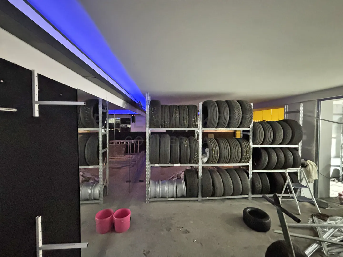 A storage room with rows of tires stacked on metal shelves, two pink buckets, and blue lighting.
