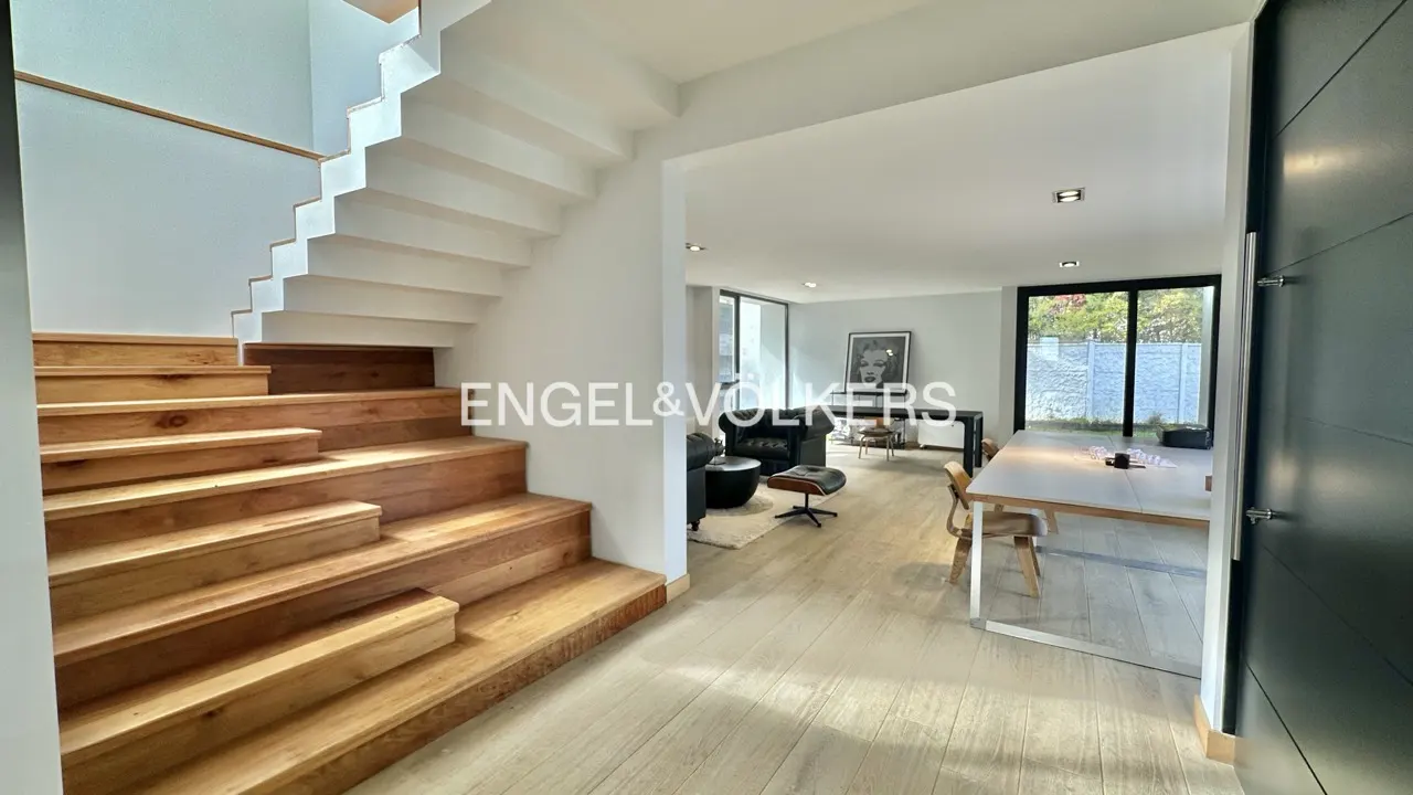 Interior view of a modern home with wooden stairs, white walls, and an open-plan living area with furniture and natural light.