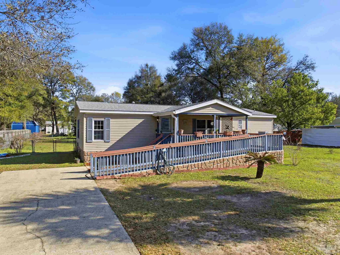 Tan single-story home with a gray roof and blue shutters. A long ramp leads to the front porch. Trees and a blue sky are in the background.