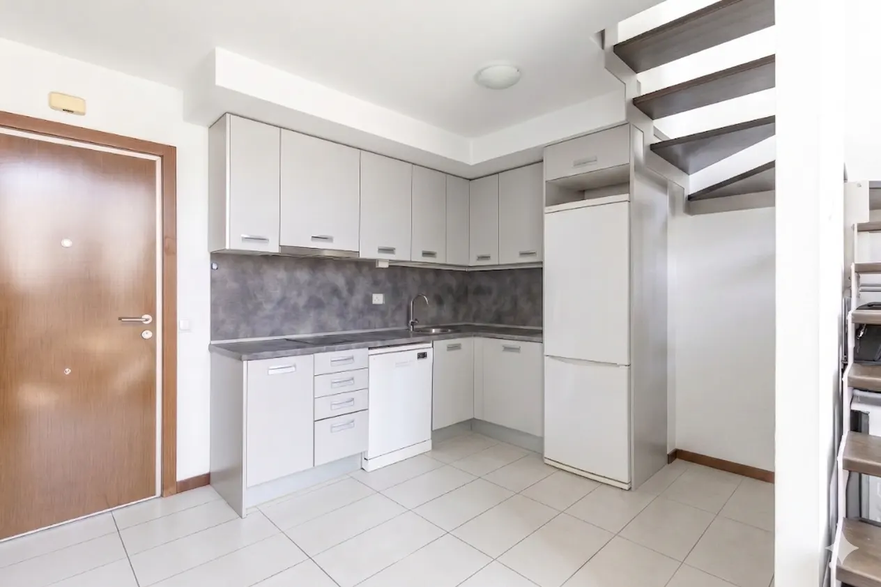 A bright kitchen with white cabinets, a gray backsplash, and a white tiled floor. A brown door is on the left, and a spiral staircase is on the right.
