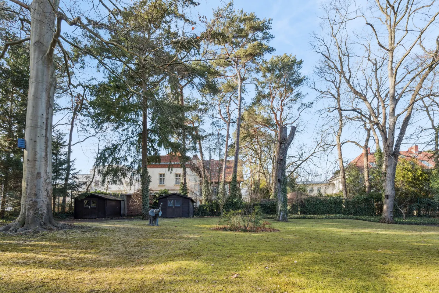 A grassy yard with trees, sheds, and an elephant statue in front of a white house with a red roof.