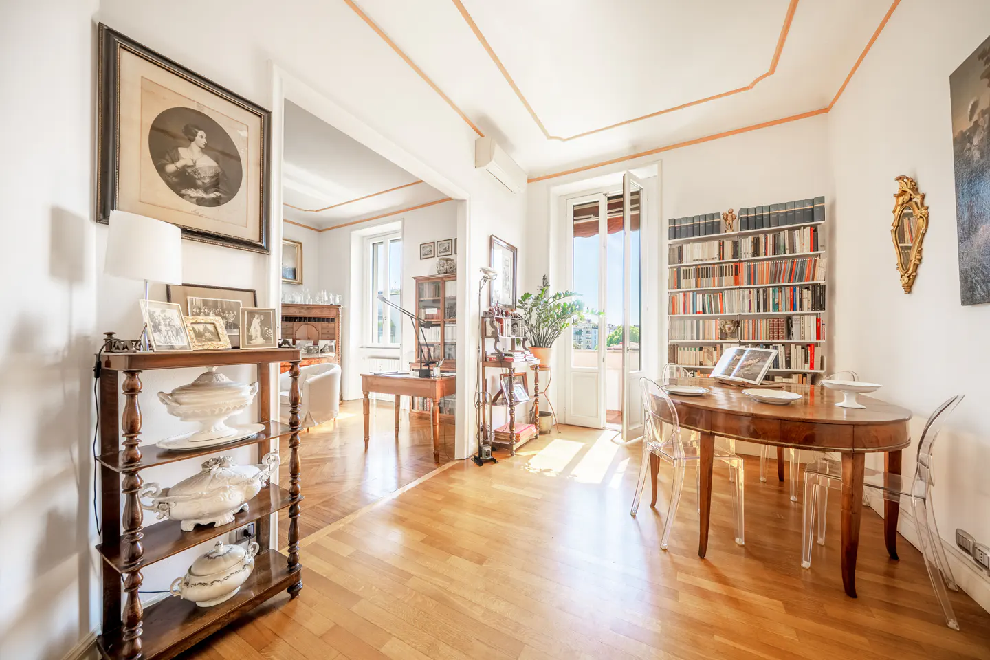 A bright, airy living room with hardwood floors, a bookshelf, and a dining table with clear chairs.
