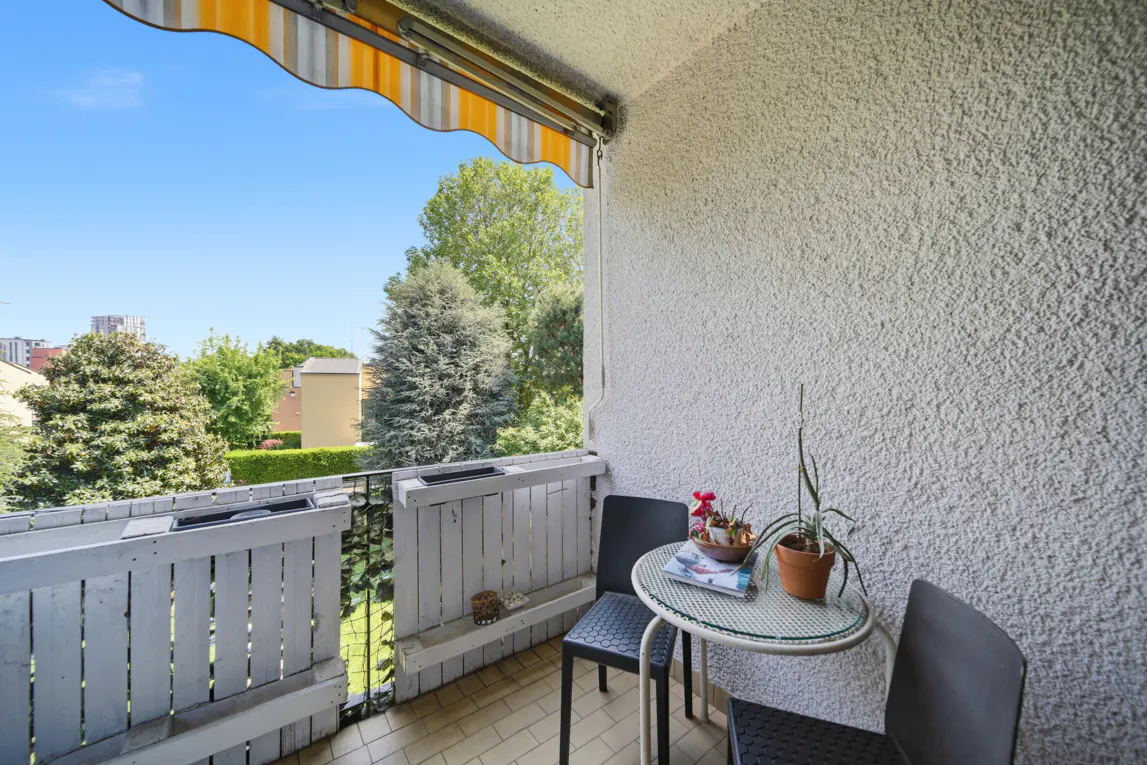 Balcony with two black chairs and a small round table with a plant and magazine. White textured wall and yellow striped awning. Trees and blue sky in background.