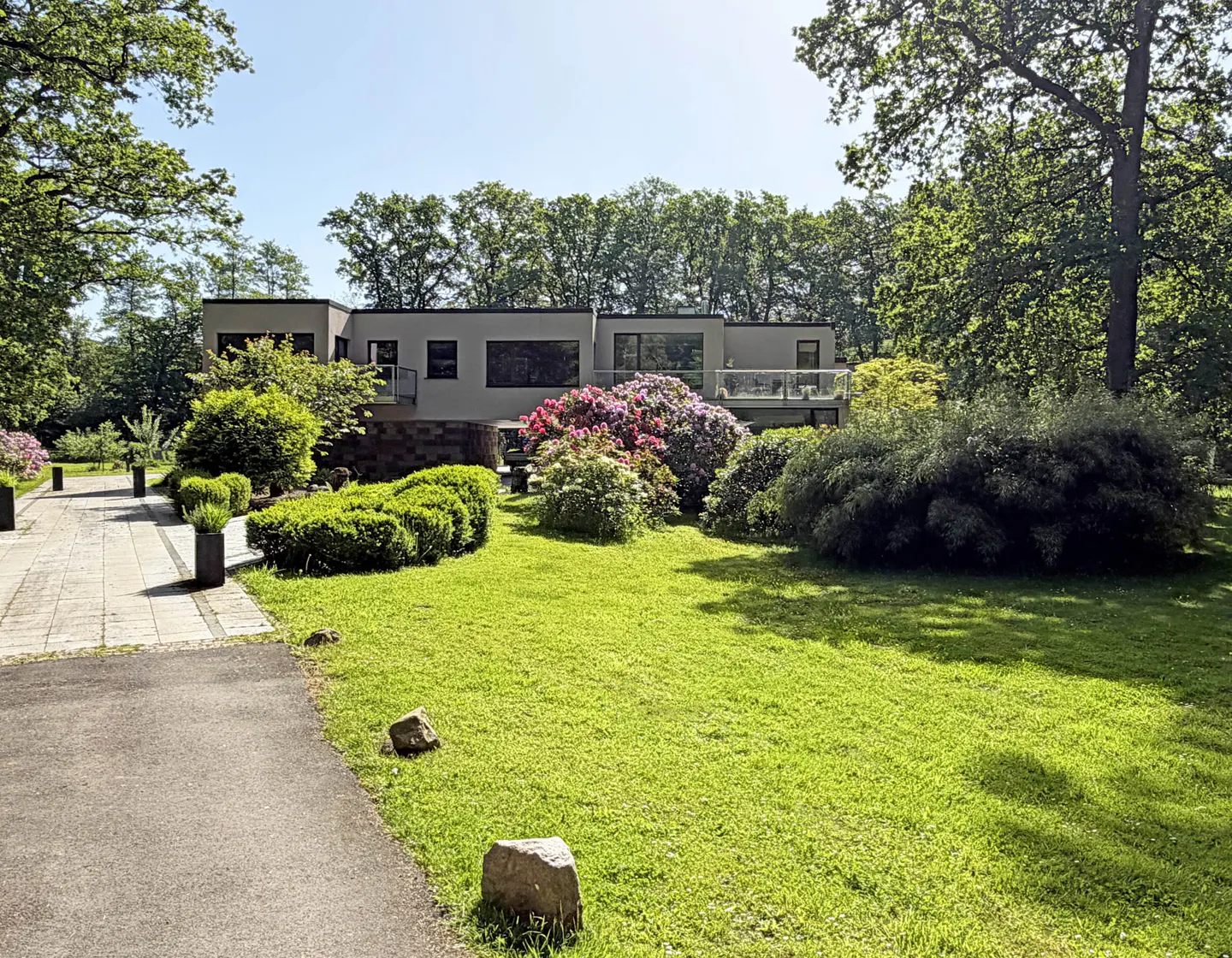 Modern house with a flat roof, stone accents, and balcony, set in a lush green garden with blooming bushes and tall trees.