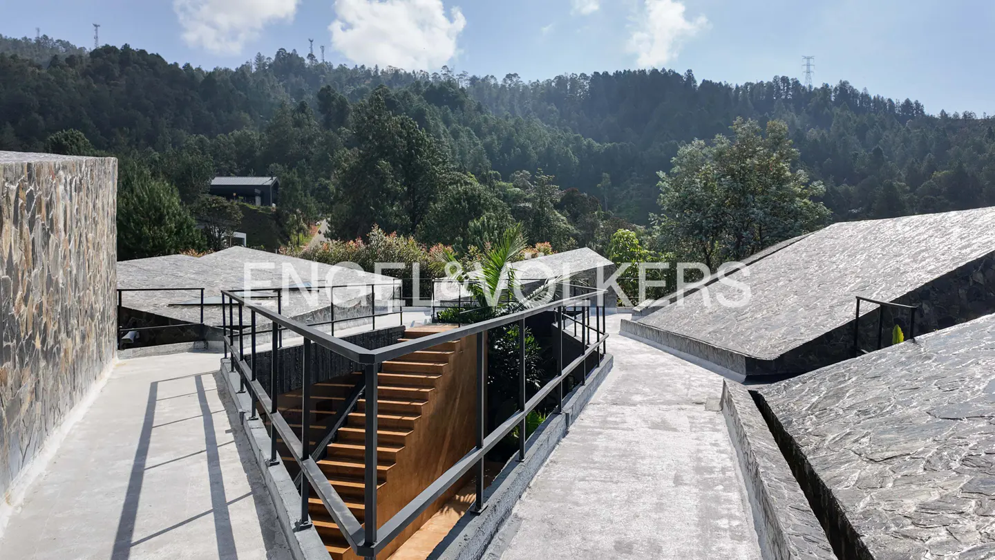 Exterior view of a modern house with stone walls, flat roofs, and black railings, set against a backdrop of lush green mountains.
