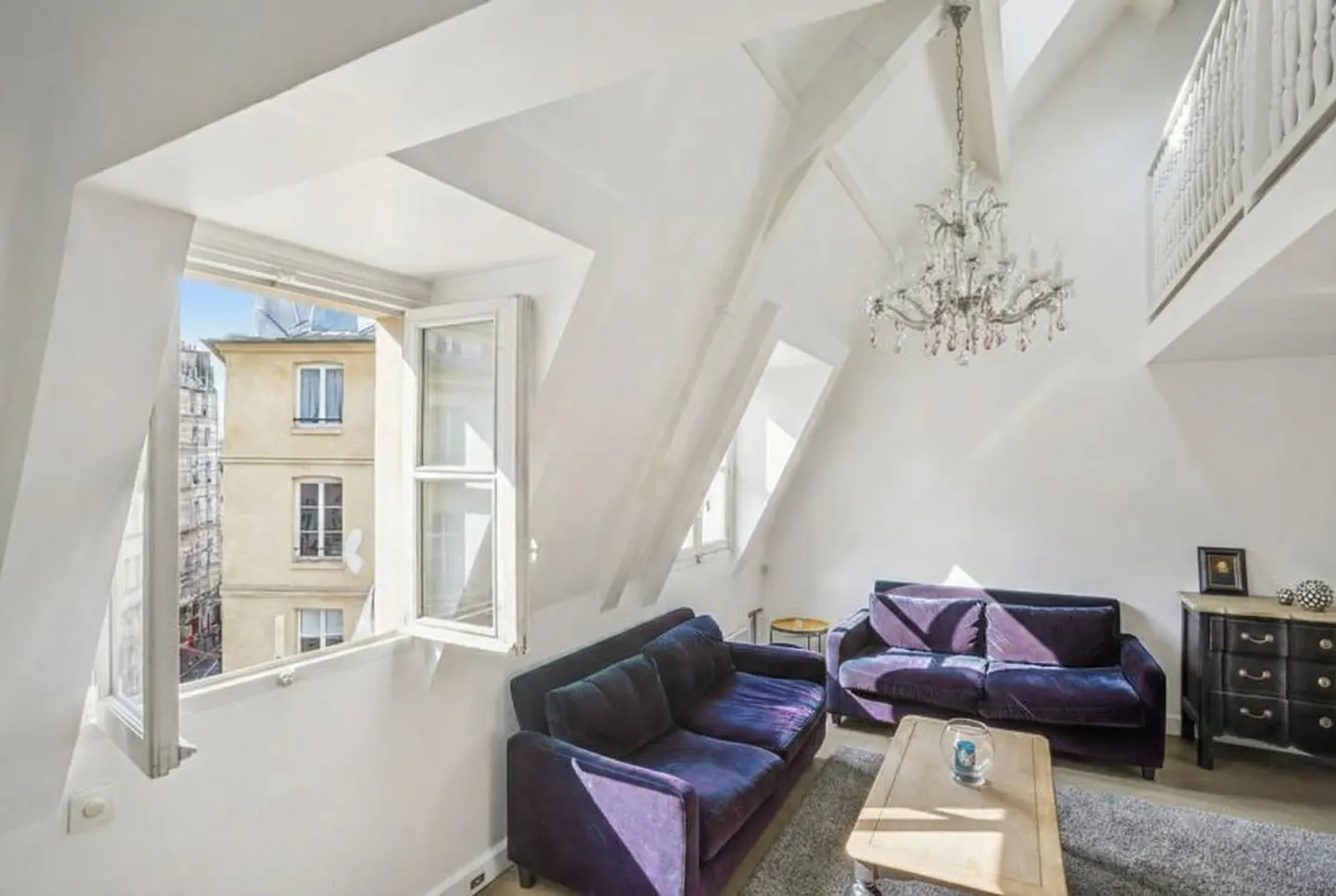 Bright attic apartment with white walls, purple sofas, and a crystal chandelier. An open window shows a building exterior.