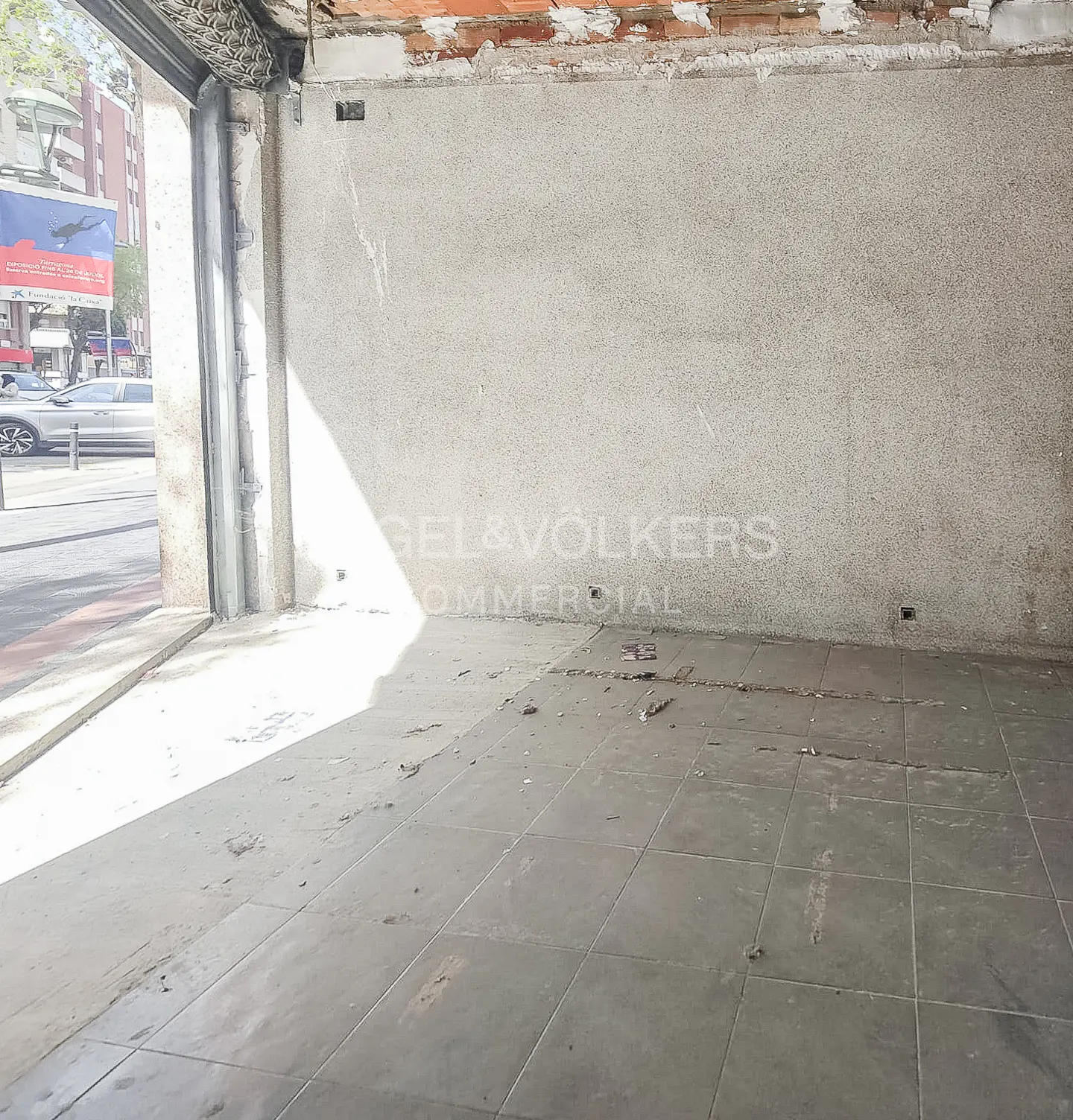 Empty commercial space with gray tiled floor, concrete walls, and open storefront to a city street.