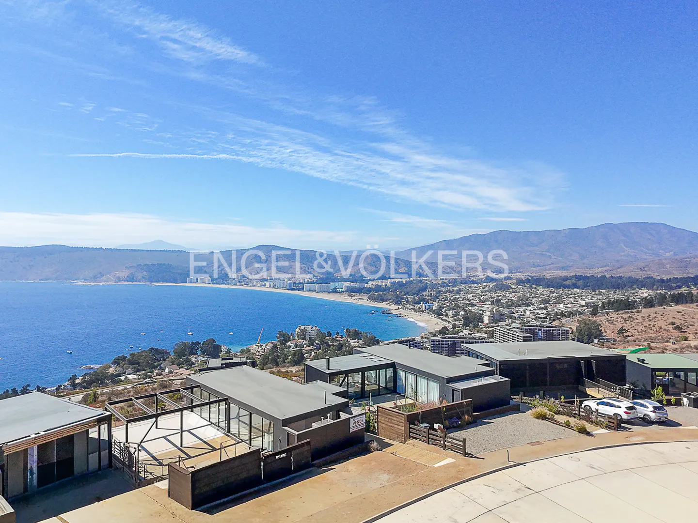 Modern home overlooking a blue ocean and beach under a bright blue sky. Two white cars are parked in front of the house.