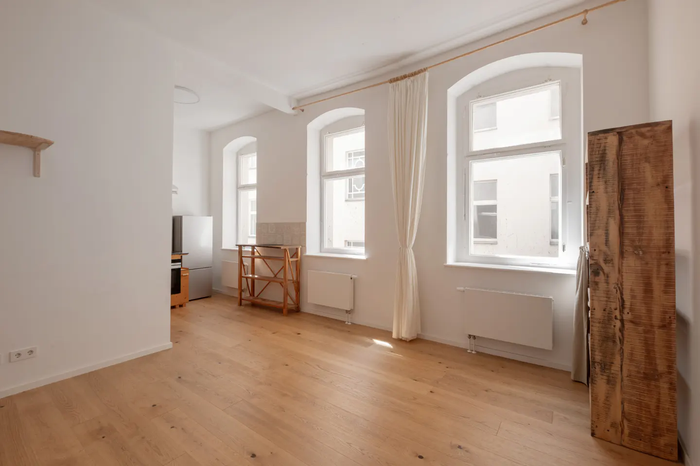 Bright room with light wood floors, white walls, arched windows with curtains, and a rustic wooden cabinet.