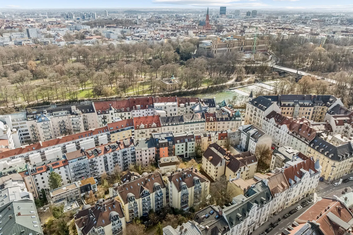 Aerial view of Munich, Germany, featuring colorful buildings with red roofs, a park with bare trees, and a river running through the city.