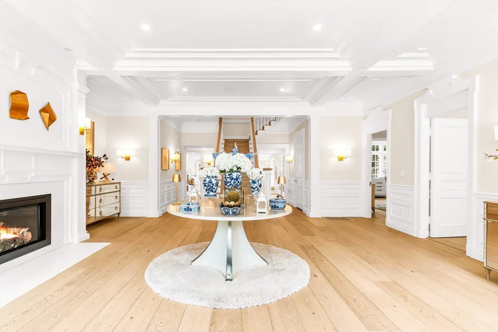 Bright foyer with wood floors, white walls, and trim. A round table with blue and white vases sits on a white rug. A fireplace is on the left.