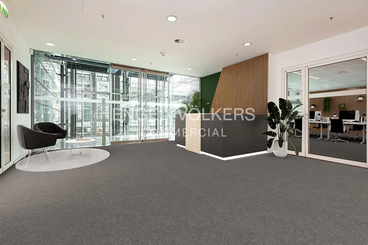 Office lobby with gray carpet, glass doors, and a black reception desk with wood accents. Two black chairs sit on a white rug.