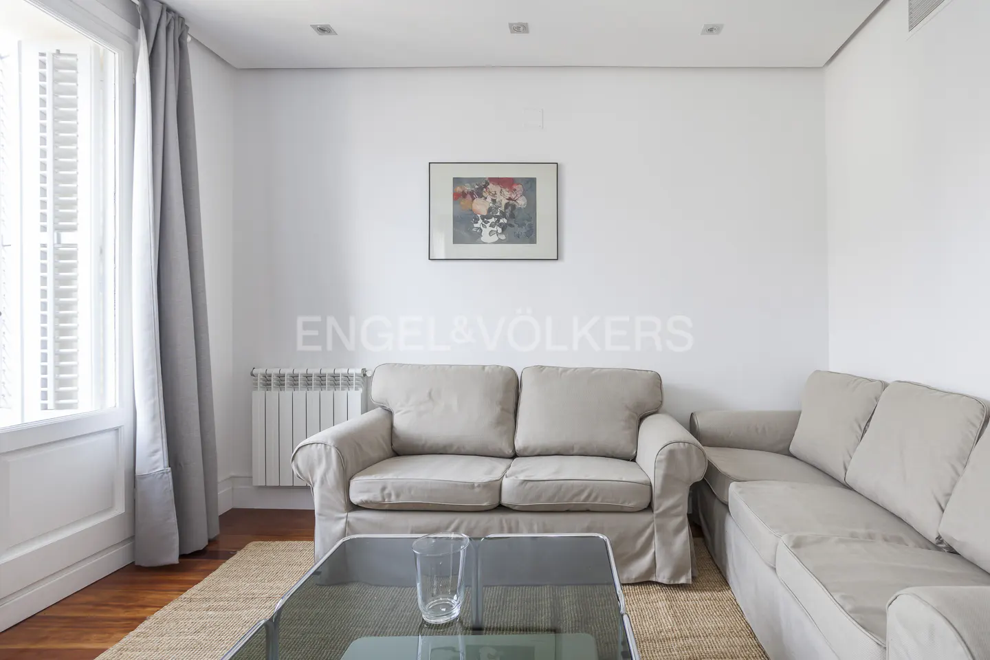 Bright living room with a beige sofa, glass coffee table, hardwood floors, and a framed abstract painting on a white wall.