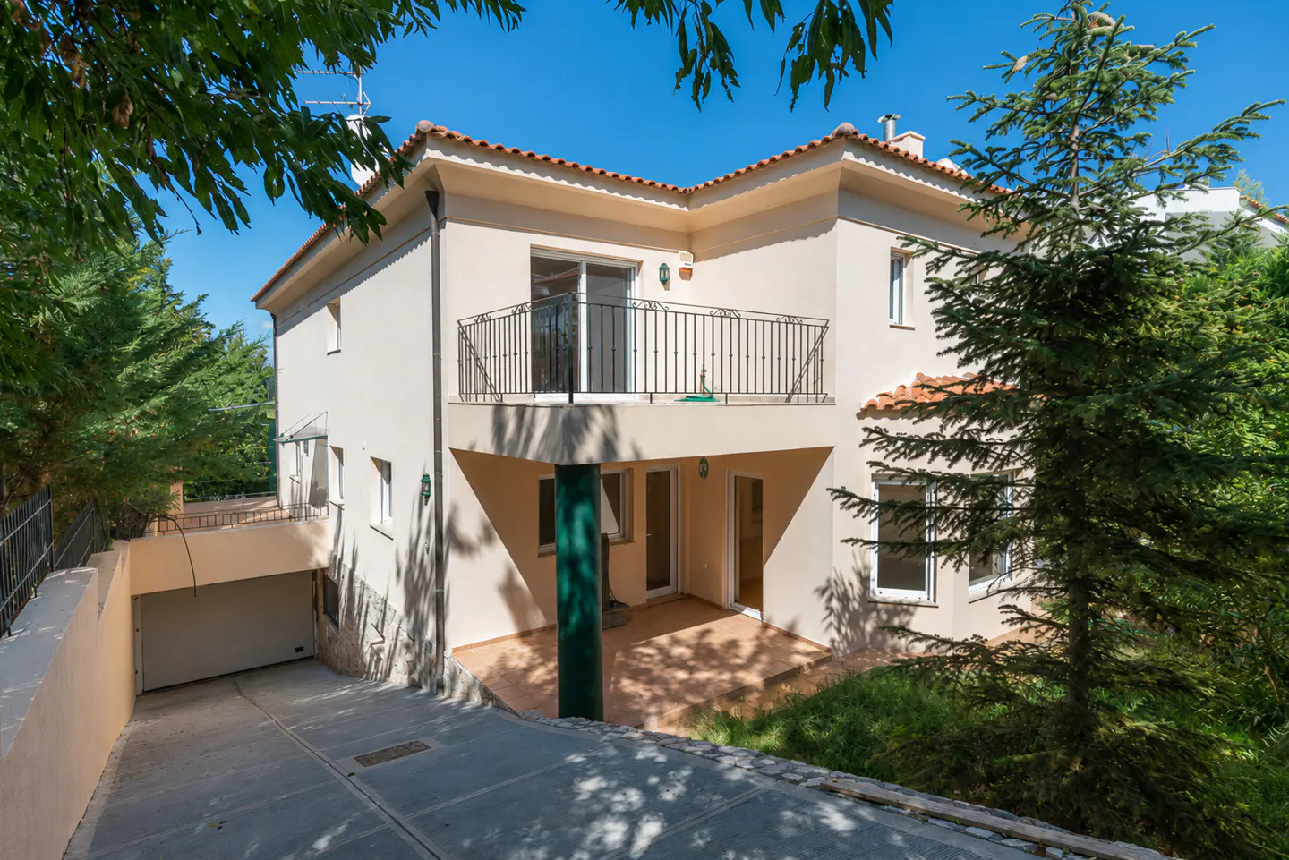 Two-story beige house with a red tile roof, black balcony railing, and a driveway leading to a garage. Green trees surround the house.