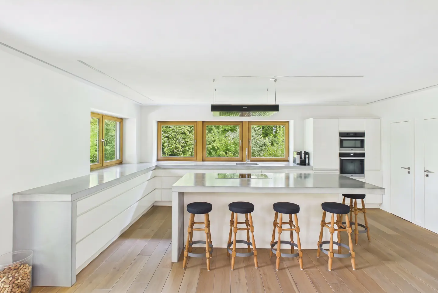Bright, modern kitchen with white cabinets, wood floors, and a large island with five stools. Windows overlook lush greenery.