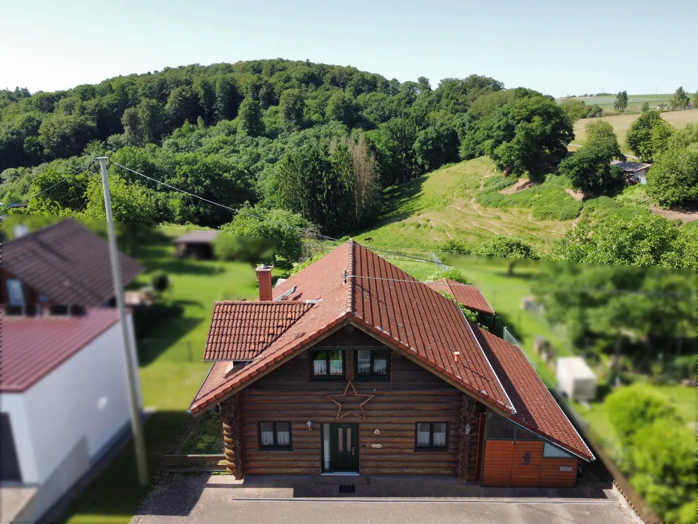 Aerial view of a rustic log cabin with a red tiled roof, featuring a star decoration, set against a lush, forested hill.
