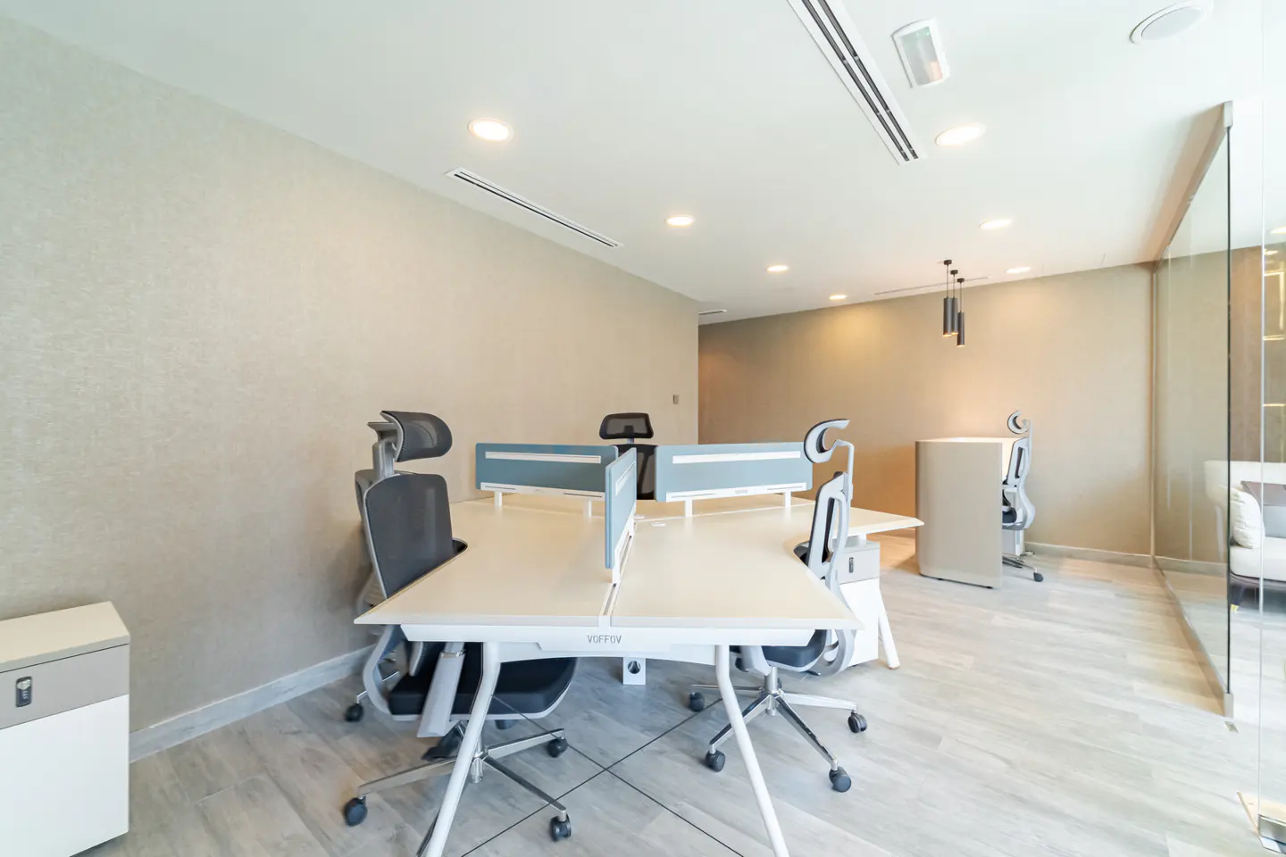 Office space with four desks, gray chairs, and light blue dividers. The walls are beige, and the floor is light gray.