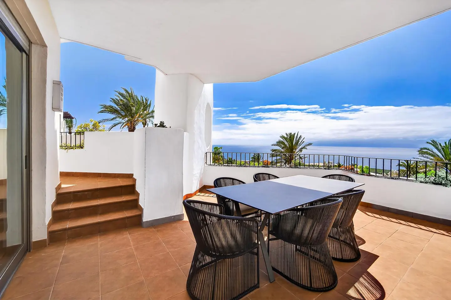Outdoor patio with a table and chairs on a tiled floor, overlooking the ocean and blue sky with palm trees.