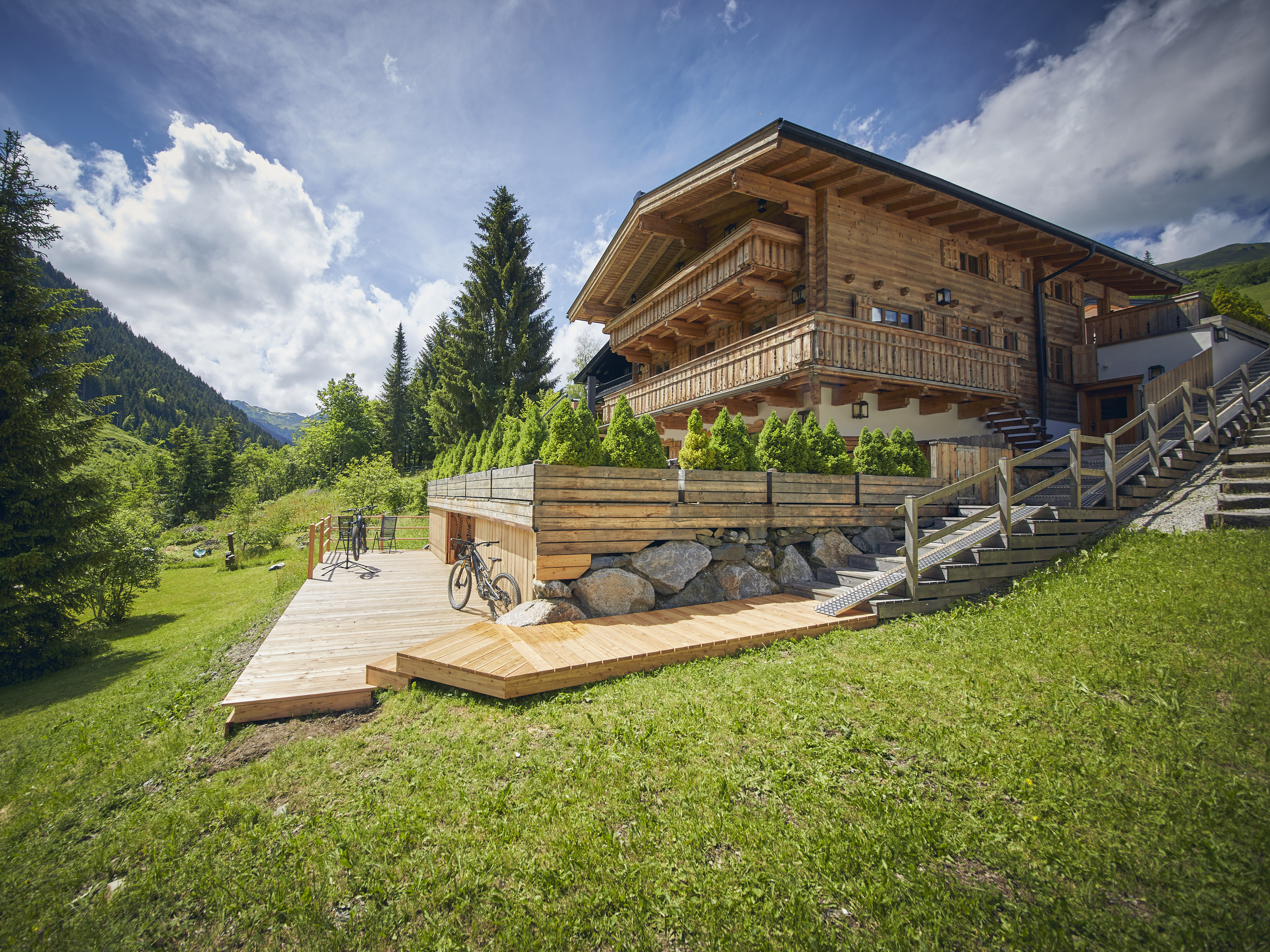 A rustic wooden chalet with a balcony, featuring a large deck with bicycles, set against a lush mountain landscape. [AI-generated text]